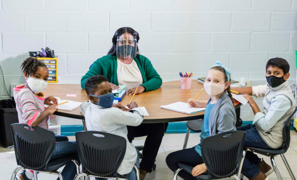 A multi-ethnic group of four children wearing face masks sitting at a table with their elementary school teacher, a mid adult African-American woman in her 30s, wearing a mask and face shield. They have returned to school during the covid-19 pandemic.